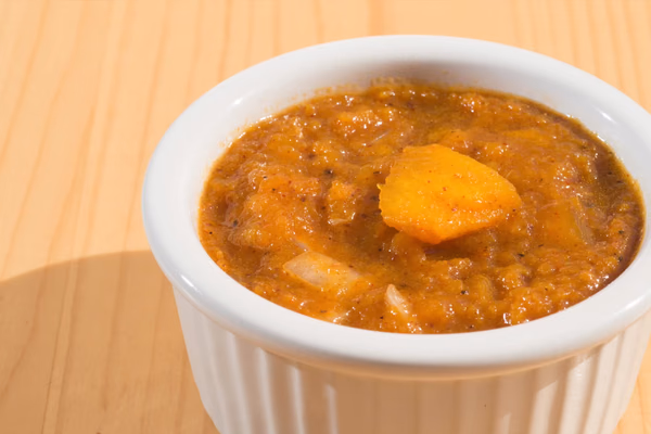 Tigela branca com sopa cremosa de cor alaranjada, vista de perto sobre uma mesa de madeira clara