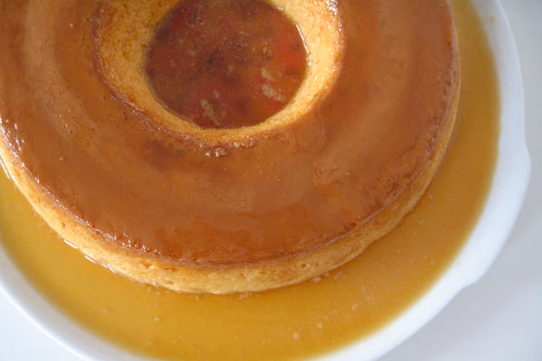 Pudim de leite condensado desenformado em prato branco, coberto por calda de caramelo