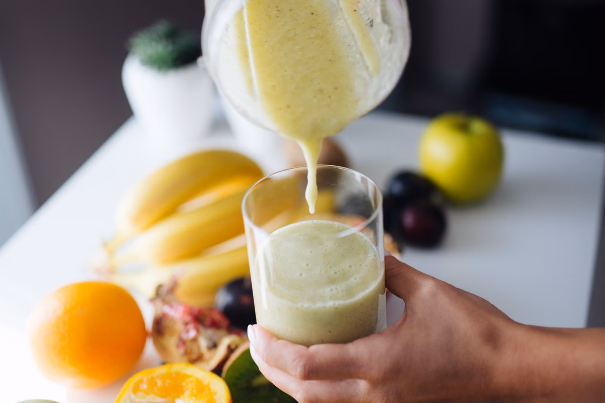 Vitamina cremosa sendo servida do liquidificador para um copo, com bananas ao lado